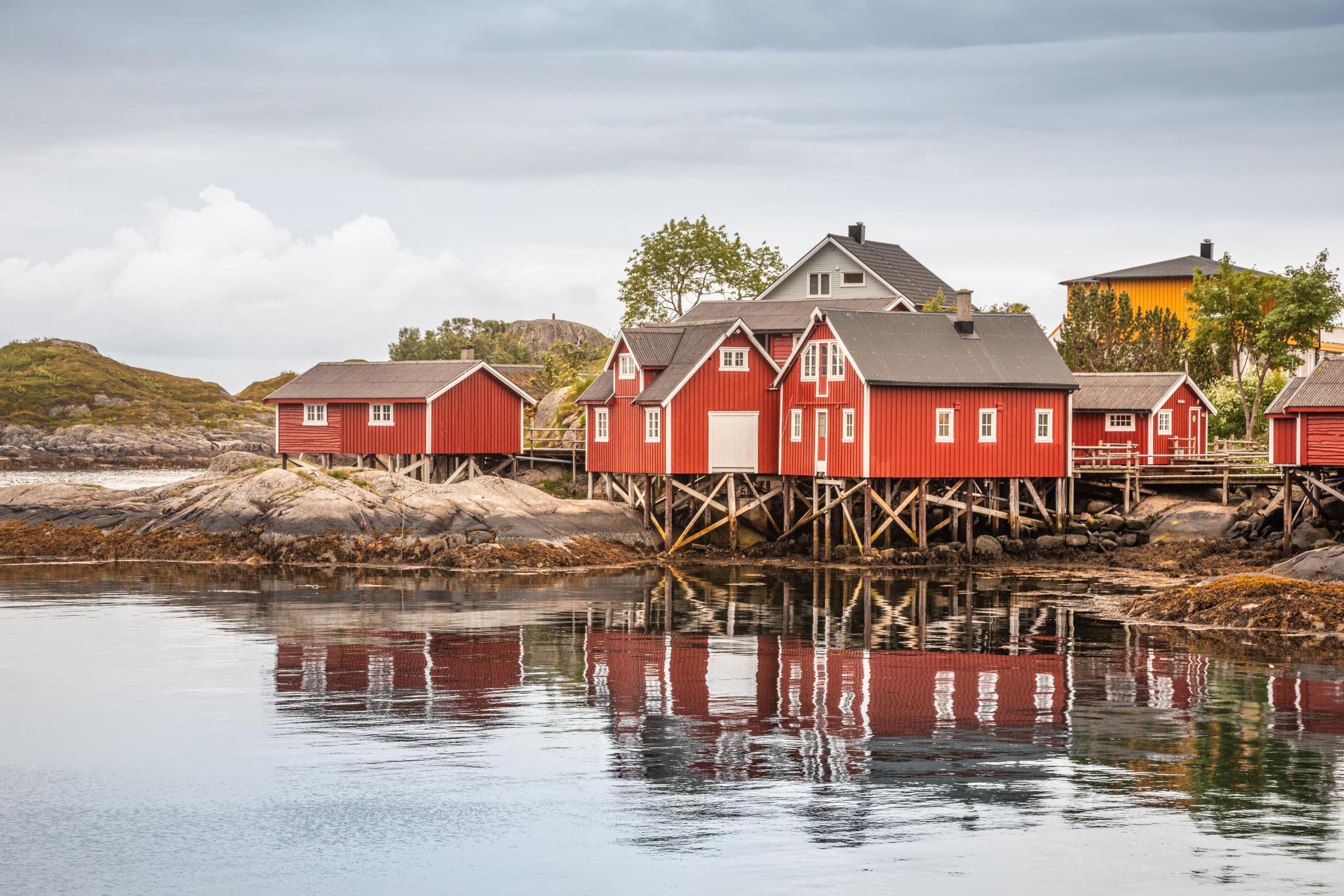 Foto Food Event in Münster – Bild der Norwegen Fjorde. Es stehen mehrere rote Stelzenhäuser aus Holz mit weißen Fenstern am Wasser. Im Hinergrund steht noch ein weißes und ein gelbes Holzhaus. vorne sieht man Wasser und die Spieglung der Häuser.