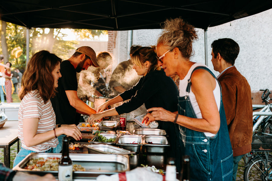 Catering in Münster mit gemeinsamen Outdoorevent. Es dampft und die Menschen lachen, unterhalten sich und kochen gemeinsam. In der Mitte stehen mehrere Behälter mit Essen.