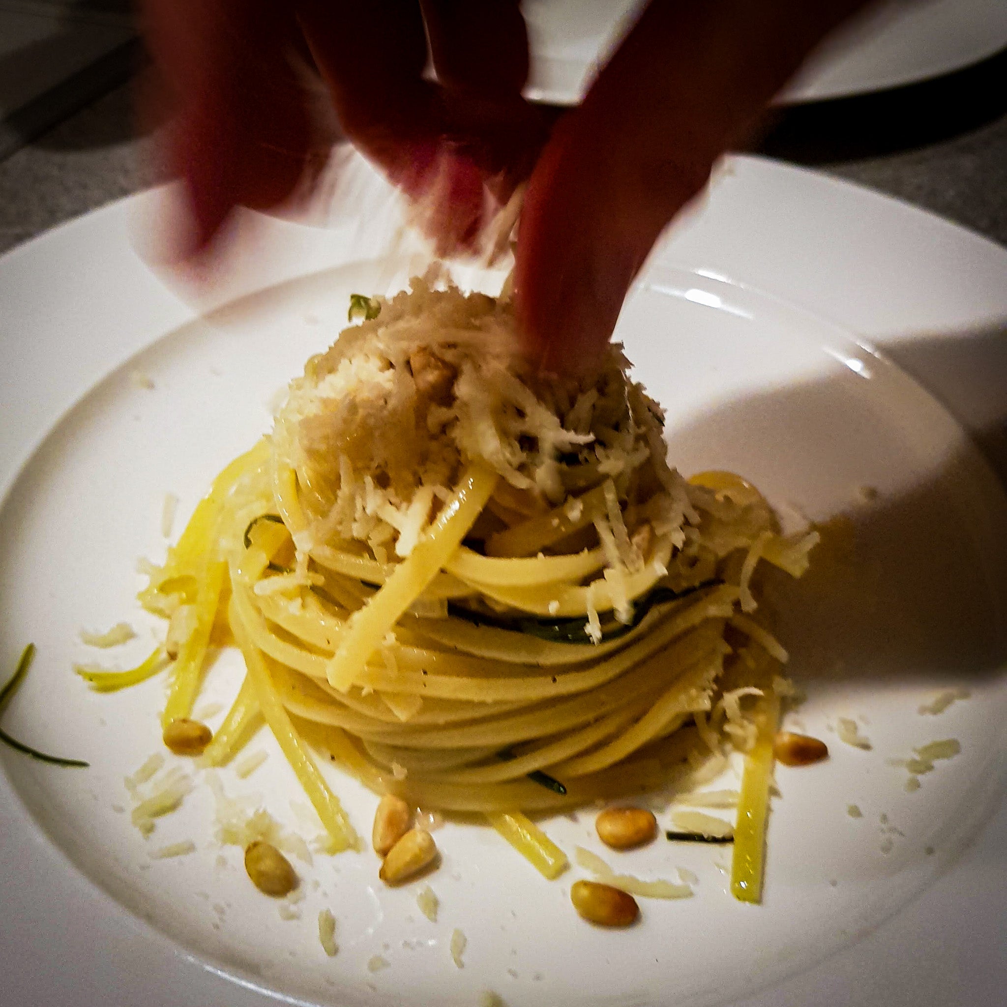 Hand in Bewegung die Parmesan auf ein Nest Nudeln mit Zucchini und Pinienkernen wirft.