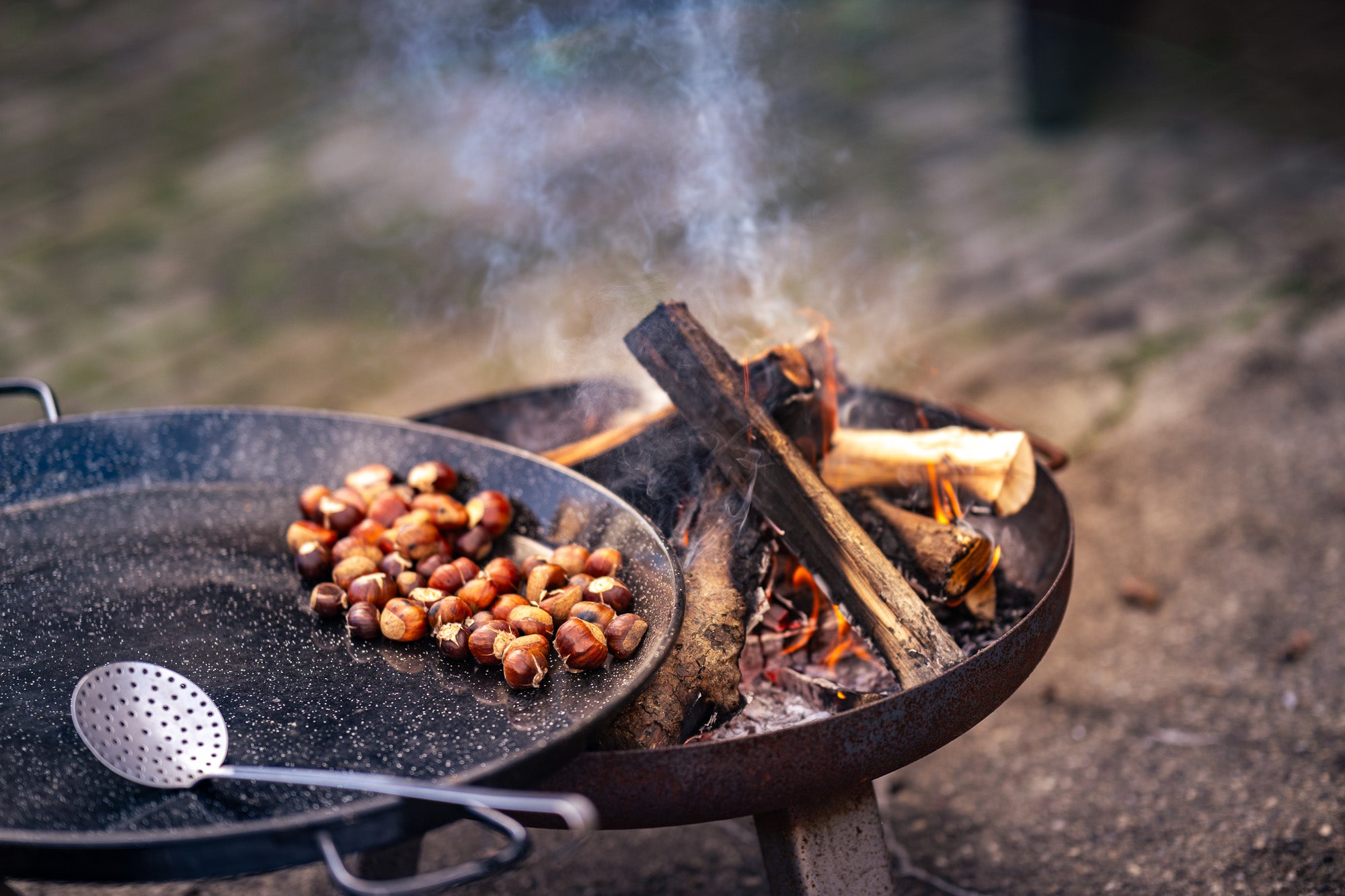 Showcooking Münster und Catering. Auf dem Bild sieht man eine Feuerschale und eine große Feuerpfanne in der Maronen gebraten werden. Vorne liegt in der Unschärfe ein silberner Schöpflöffel. Das Feuer raucht.