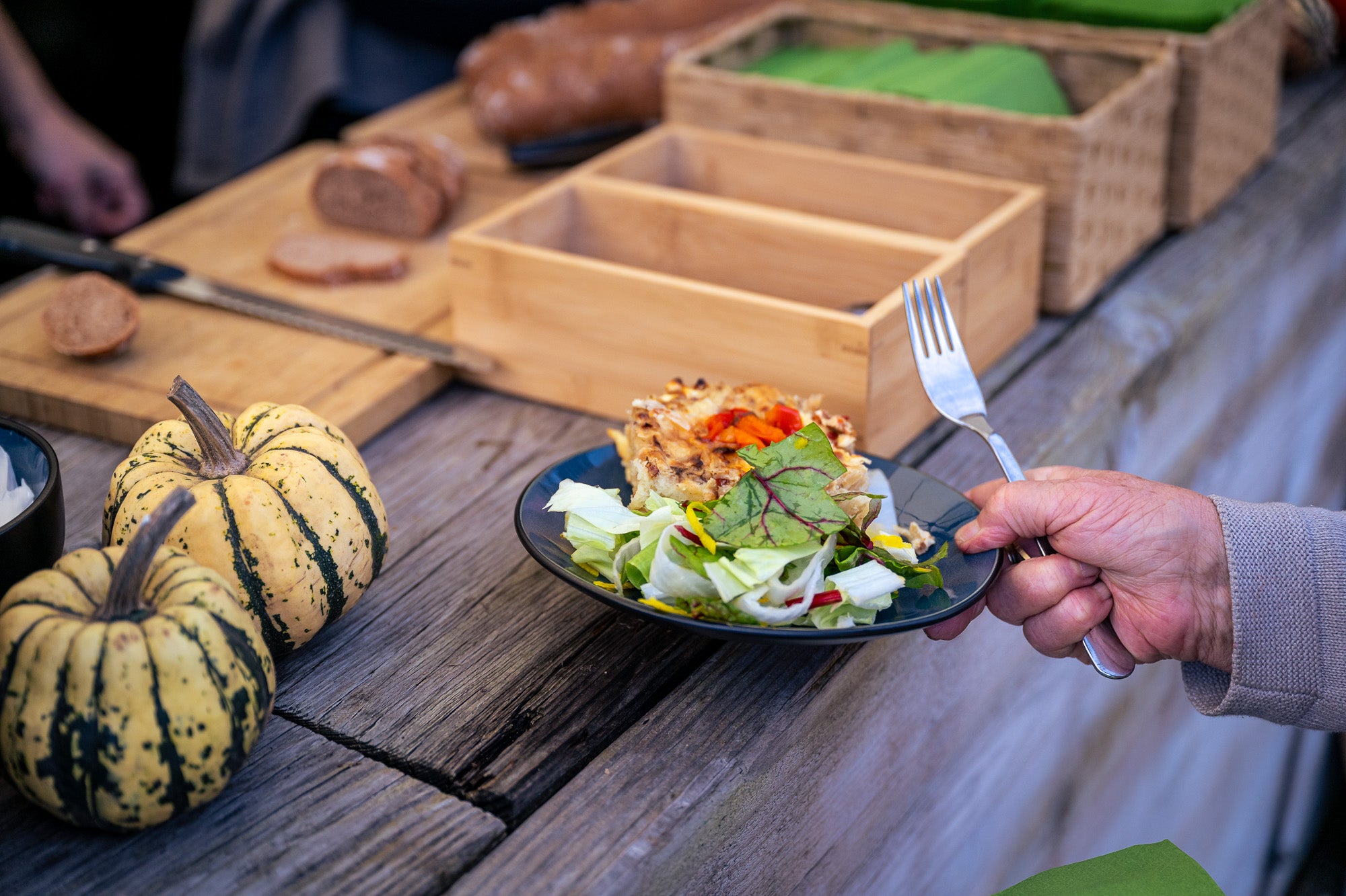 Catering in Münster: eine Hand hält einen Teller mit frischem, günem Salat und Zwiebelkuchen, wie auch eine Gabel über einer Holztheke. Links stehen frische Kürbisse. Im Hintergrund steht frisches geschnittenes Brot. Man sieht im Anschnitt noch eine andere Hand auf der anderen Seite der Theke. Es stehen noch Besteck und Serviettenkästen auf der Holztheke.