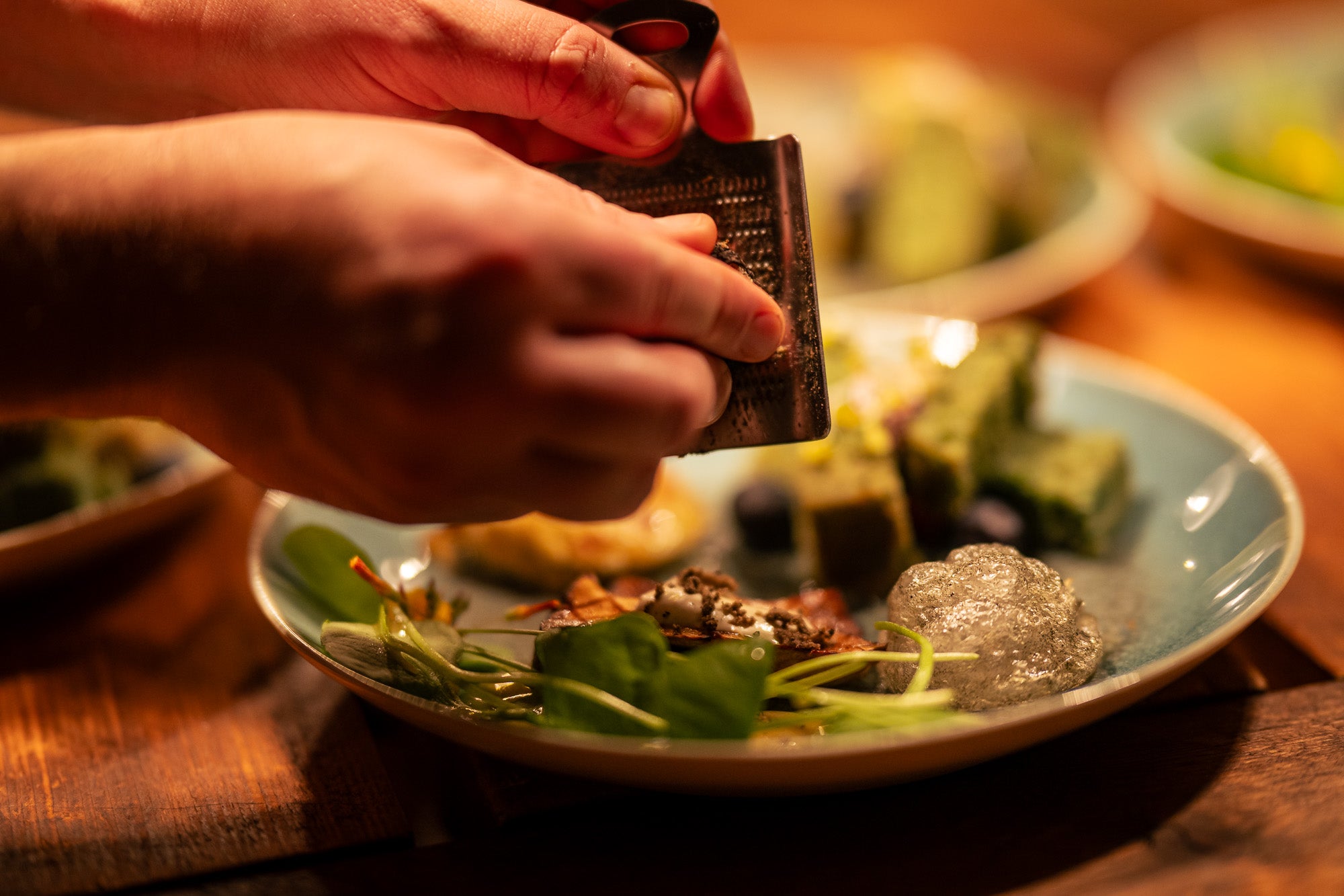 Ein Teller mit Essen auf einem Holztisch. Im Hintergrund weitere Teller. Im Vordergrund Zwei Hände die eine Reibe nutzen. 
Bildrecht bei Thomas Mohn.