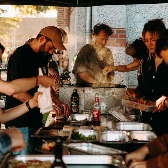 Foodbike in Münster - Catering Outdoor – Elias Eckel kocht bei einem Outdoorevent in einer großen Pfanne, während andere Menschen mitkochen und Gemüse schneiden und Essen vorbereiten.