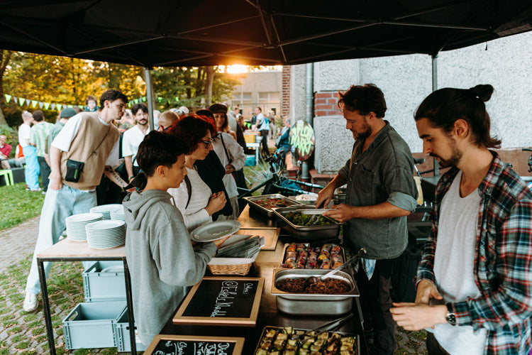 Foodbike in Münster - Catering Outdoor – Ein Cateringstand steht bei einem Outdoor Sommerfest. Elias Eckel und ein Mitarbeiter servieren den anstehenden Menschen unterschiedliches Essen, welches in GN-Behältern vorbereitet und vorher gemeinsam gekocht wurde. Es steht eine Schlange an Menschen davor. Im Hintergrund sieht man noch ein bisschen Sonnenlicht