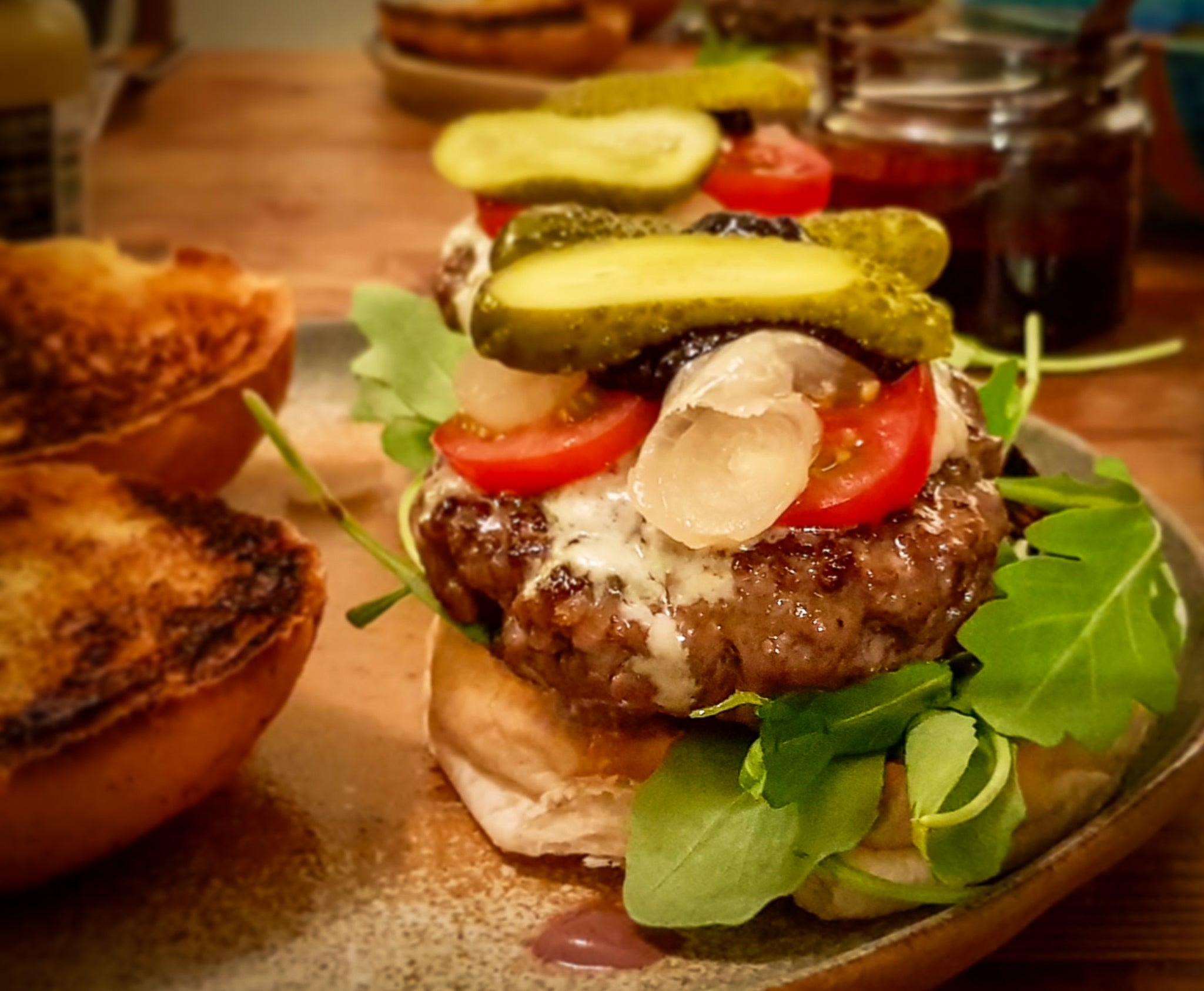 offener Burger mit Essiggurken, Tomaten, Rucola und Lardo. Im Hintergrund Saucen und Teller.