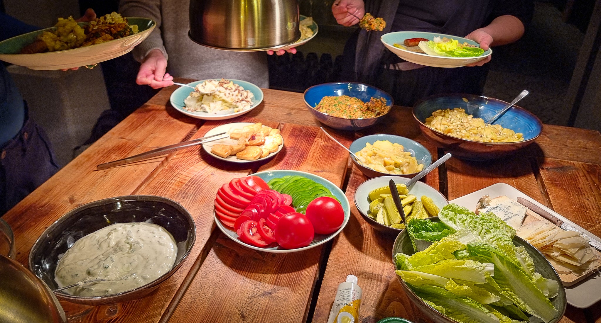 Holztisch mit Schüsseln und diversen Gemüse-Zutaten. Menschen außenherum die sich bedienen.