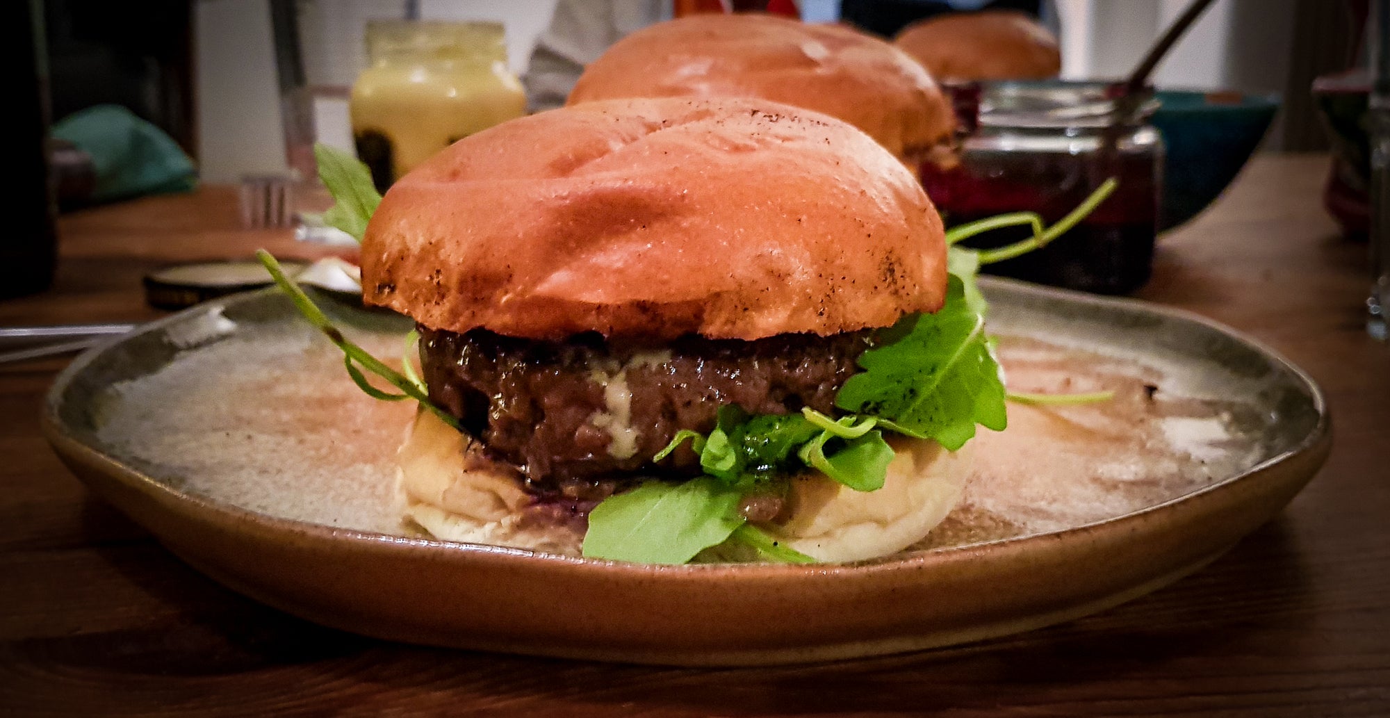 Burger mit Rucola in der Seitansicht. Im Hintergrund Saucen und Schüsseln.