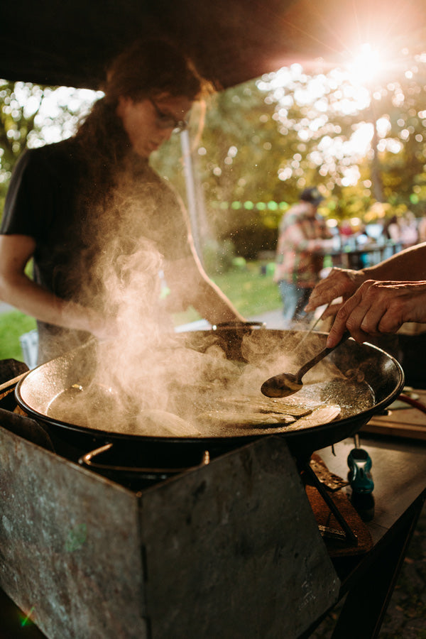 Showcooking Outdoor in Münster - Foodbike- Fahrrad Catering. Bei einem Outdoorevent braten Hände in einer großen Pfanne Zucchini Streifen an. auS der Pfanne dampft es. Im Hintergrund steht ein Mann in der unschärfe, der ebenfalls etwas kocht.