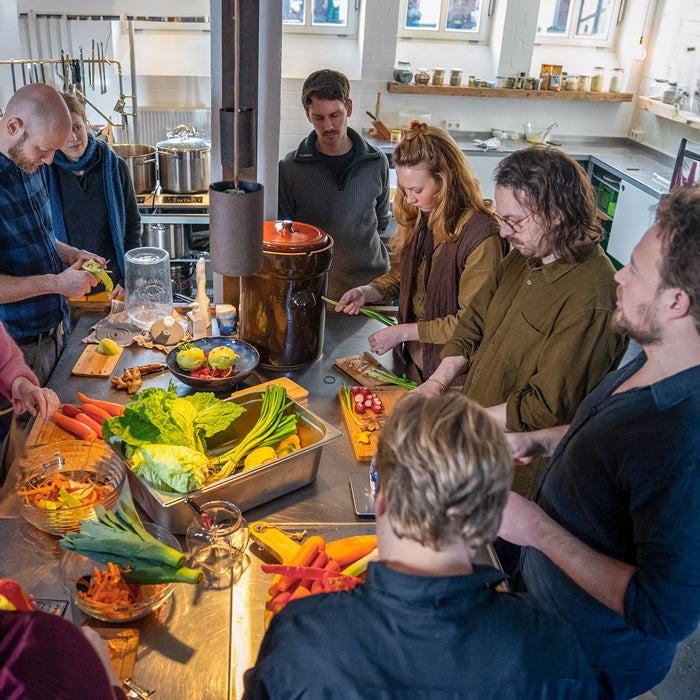 Kochkurs mit mehreren Menschen in Münster – mehrere Personen kochen gemeinsam in der Kochwerkstatt von Elias Eckel. In der Mitte steht unterschiedliches Gemüse in verschiedenen Behältern. 