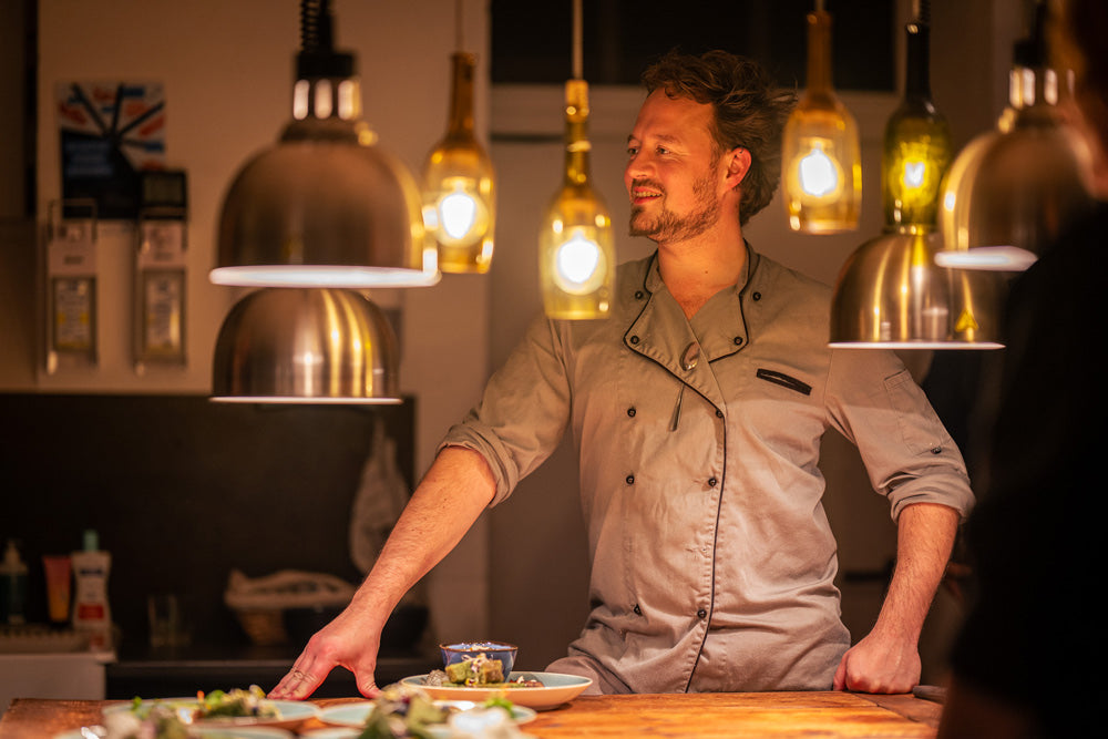 Elias Eckel in der Kochwerkstatt der Essenz. Im Vordergrund sind unscharf mehrere Lampen. Elias steht mittig im Bild an einer Kochinsel mit Essen auf der Insel und sieht nach links.  Im Hintergrund sieht man ein Wachbecken.