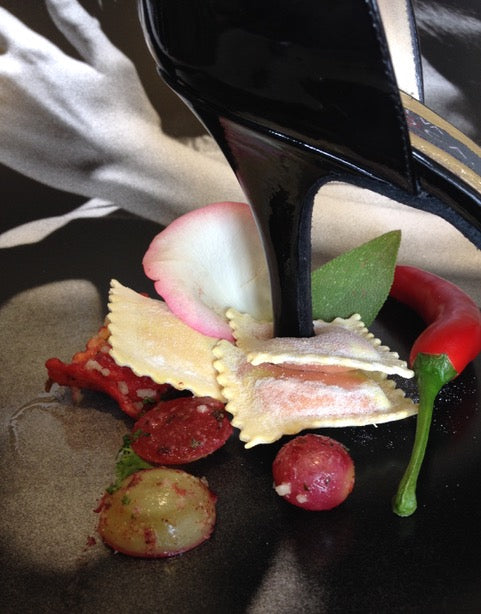 Ravioli, Tomaten und Chili auf einem Teller. Die Ravioli sind von dem Absatz eines hochhackigen Schushs aufgespießt. 