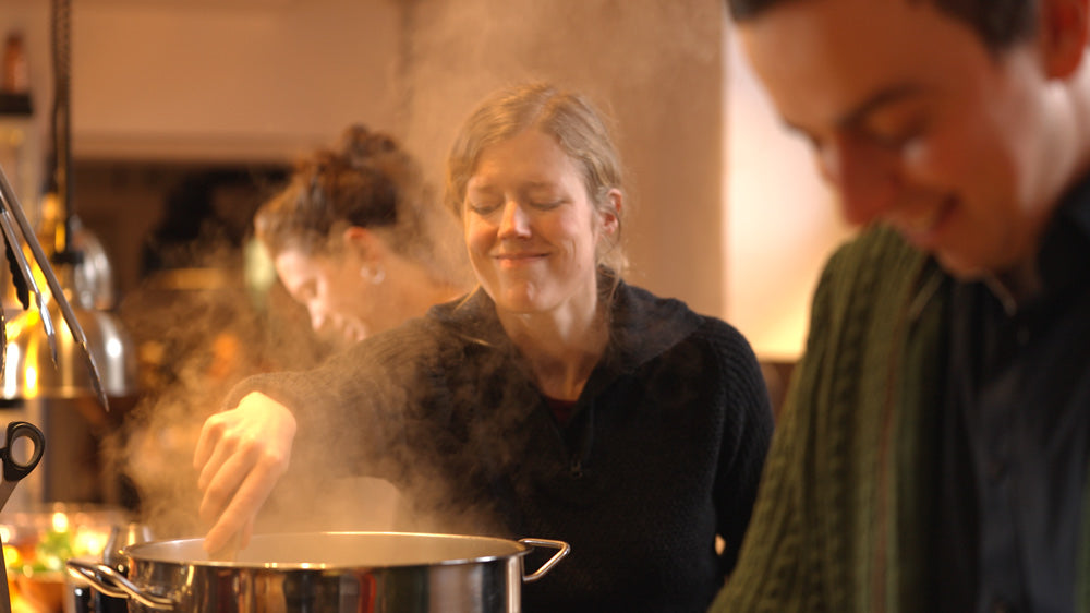 Kochkurse in Münster - Workshop kochen Eine Frau mittleren Alters und Zopf lächelt und schaut in einen Kochtopf in dem sie gerade etwas umrührt. Aus dem Kochtopf dampft es. Im Vordergrund und im Hintergrund sind Personen in der Unschärfe