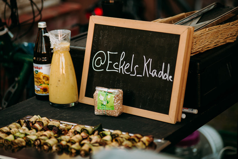  Tafel mit dem Namen Eckels_Kladde auf einem Outdoor- Event mit Catering auf einem Tisch mit Essen und Saaten, wie auch Smoothie und Öl.